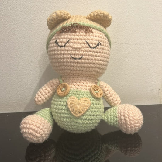A handmade crocheted doll with a beige face and limbs, wearing a light green outfit with a heart-shaped patch on the chest, sitting on a reflective black surface against a plain white wall.