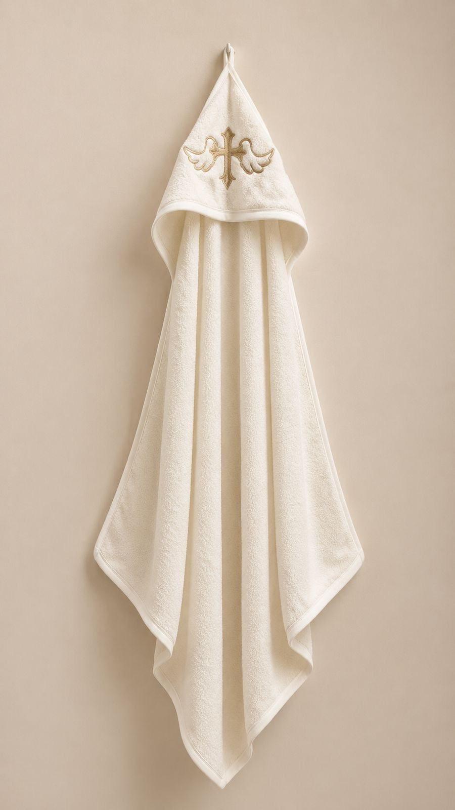 An off-white hooded towel with a gold embroidered cross and angel motif on the hood, hanging against a solid beige background.