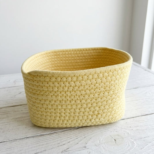 A light yellow crocheted storage basket with a textured honeycomb pattern, sitting on a white-painted wooden surface against a white wall.
