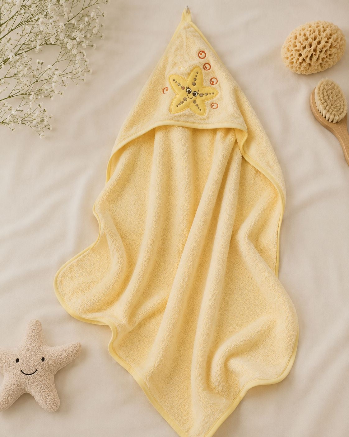A pale yellow hooded towel laid flat on a white fabric surface, decorated with a starfish motif on the hood. A small plush starfish toy, a sponge, and a wooden brush are arranged around it as styled props.