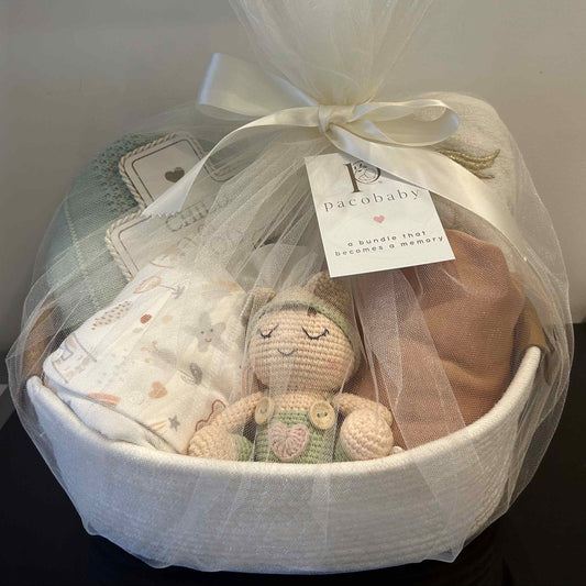 A baby gift basket wrapped in white tulle and tied with a satin ribbon, containing a crocheted doll, folded blankets, and a diaper bag. A hanging tag reads 'pacababy'.