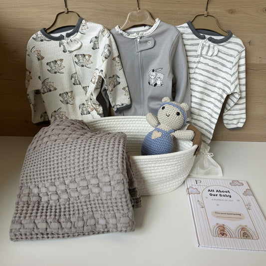 A baby clothing and accessory set displayed on a white surface, including three long-sleeve baby tops in cream, gray, and white striped patterns, a folded textured gray blanket, a white woven basket holding a blue crocheted doll, and a booklet titled 'All About Our Baby'.
