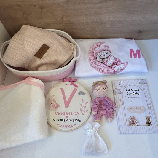 A baby gift set arranged on a white surface against a light wood background, including a pink and cream basket with a folded towel, a white cloth with a pink bunny illustration and letter M, a cream towel, a round embroidered monogram patch reading Veronica, a small pink knitted doll, and a printed card.