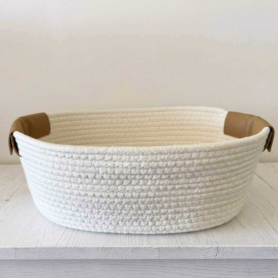 A round woven rope basket in off-white with tan leather handles, resting on a white wooden table against a plain light wall.