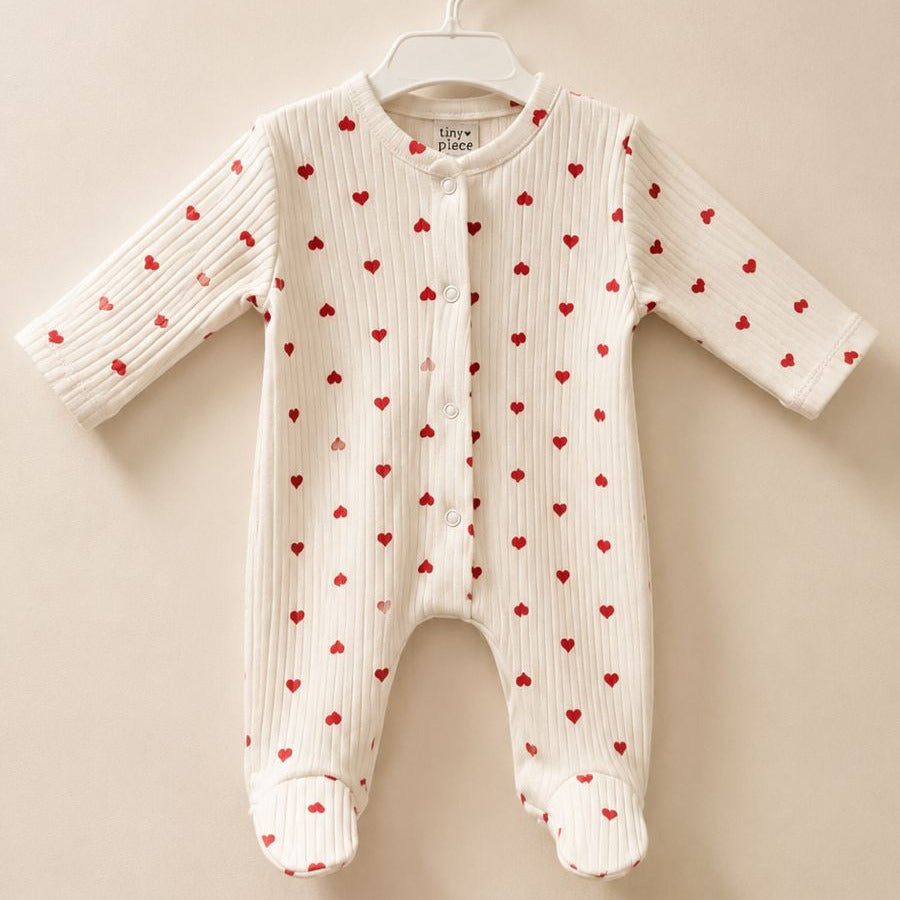 A white ribbed knit baby footed sleeper with a red heart pattern, hanging on a white plastic hanger against a solid beige background.