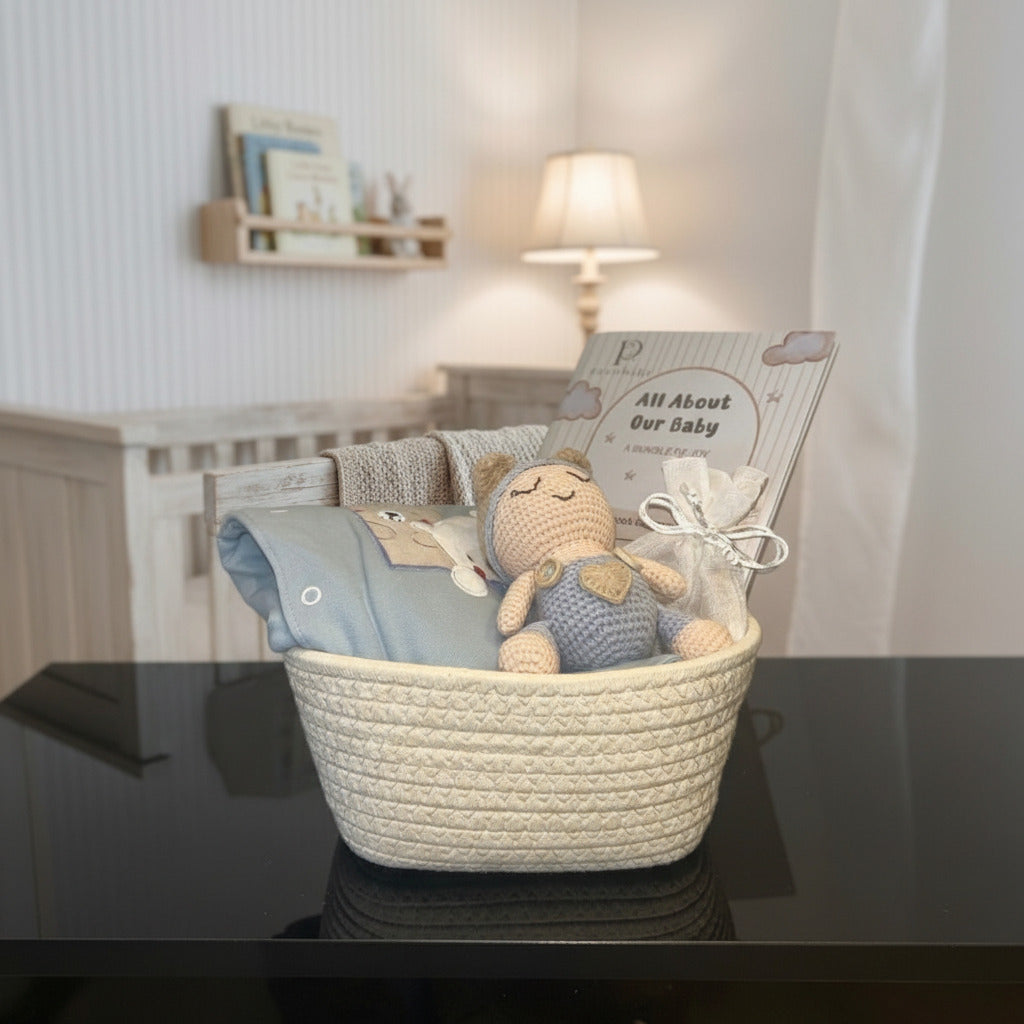 A baby gift basket set in a nursery, featuring a cream woven basket containing a blue knit blanket, a crocheted teddy bear, and a gift bag with a card that reads 'All About Our Baby'.