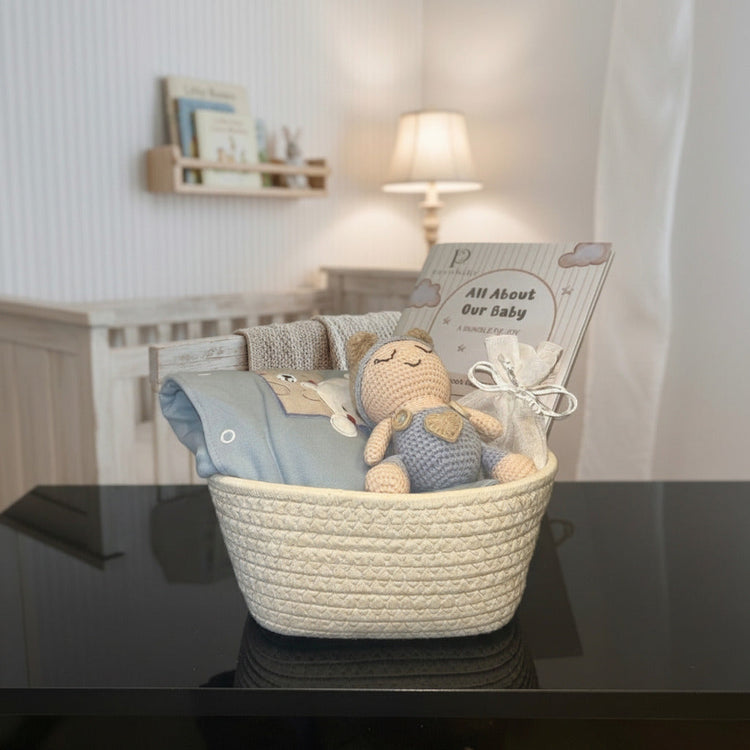 A baby gift basket set in a nursery, featuring a cream woven basket containing a blue knit blanket, a crocheted teddy bear, and a gift bag with a card that reads 'All About Our Baby'.