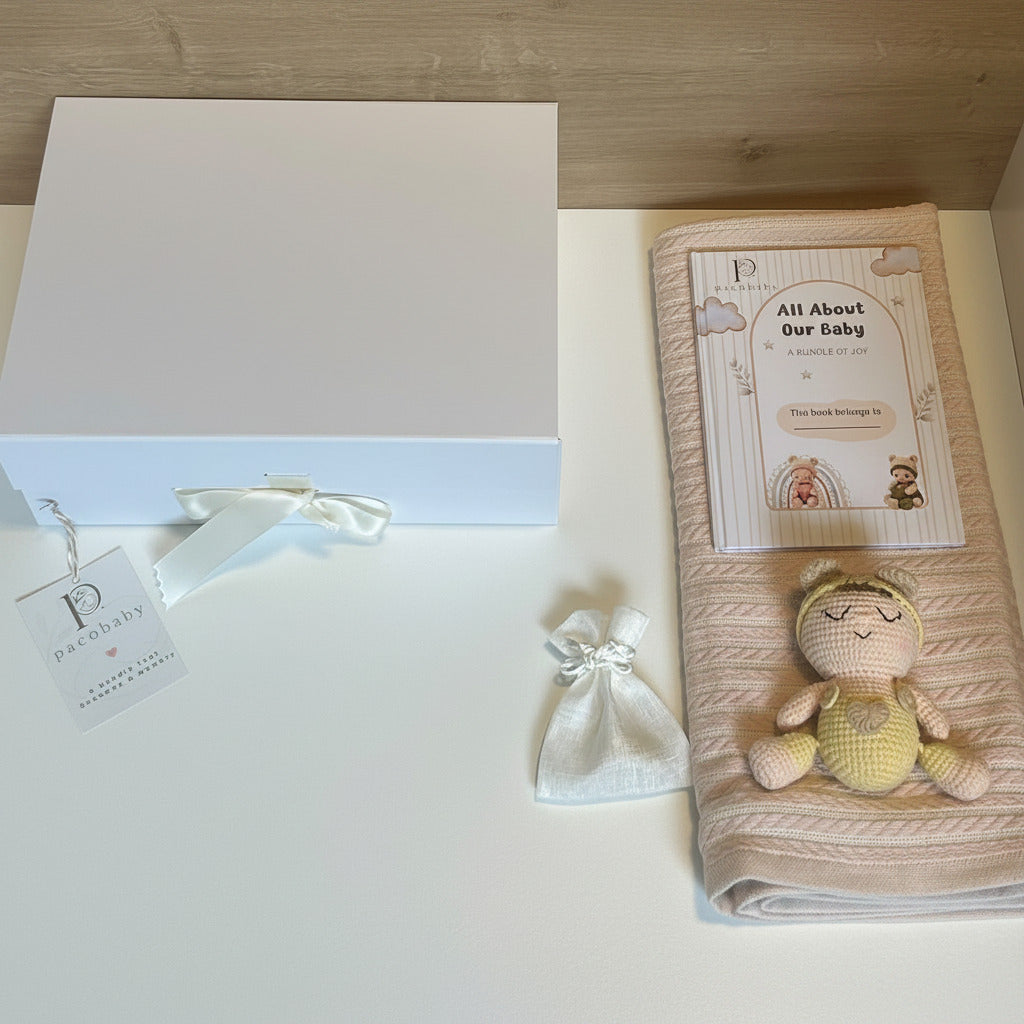 A baby gift set featuring a white box with a white ribbon and tag, a small white drawstring pouch, a folded pale pink muslin blanket with a printed card, and a crocheted yellow and beige bunny toy on a white surface.
