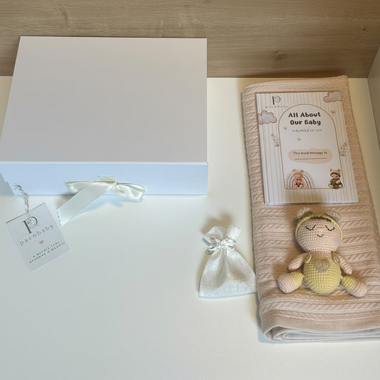 A baby gift set featuring a white box with a white ribbon and tag, a small white drawstring pouch, a folded pale pink muslin blanket with a printed card, and a crocheted yellow and beige bunny toy on a white surface.