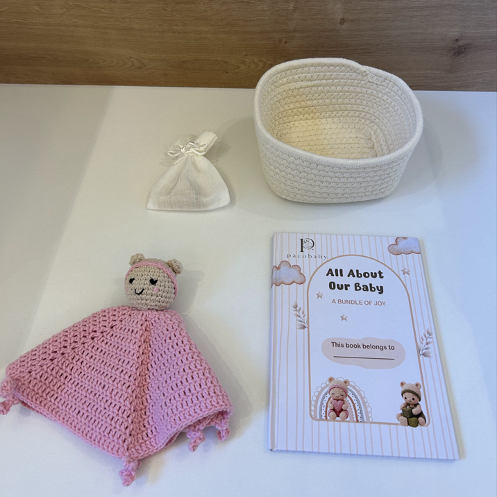 A baby gift bundle set including a pink knitted swaddle with a bear plush, a cream woven basket, a small cream bow, and a printed booklet titled 'All About Our Baby' on a white surface with a wood background.