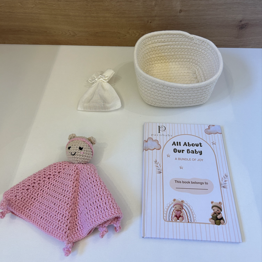 A baby gift bundle set including a pink knitted swaddle with a bear plush, a cream woven basket, a small cream bow, and a printed booklet titled 'All About Our Baby' on a white surface with a wood background.