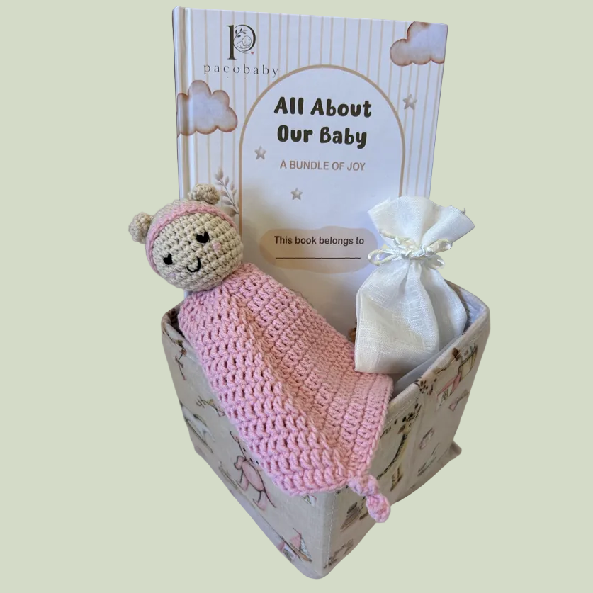 A baby gift set in a floral-patterned box featuring a pink crocheted lovey, a white drawstring bag, and a book titled 'All About Our Baby' by Pristine Baby, displayed on a white surface.