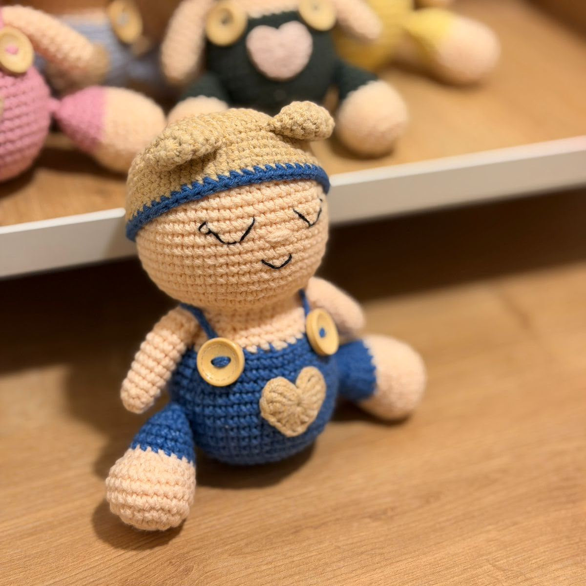 A crocheted doll in the foreground wearing blue overalls with a beige heart patch and yellow buttons, sitting on a wooden surface. Other crocheted dolls are blurred in the background on shelves.