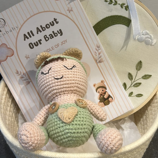 A handmade crochet baby gift set in a woven basket, featuring a cream-colored crochet doll with a green belly, a printed card reading 'All About Our Baby', and a decorated round cloth with a white ribbon bow.