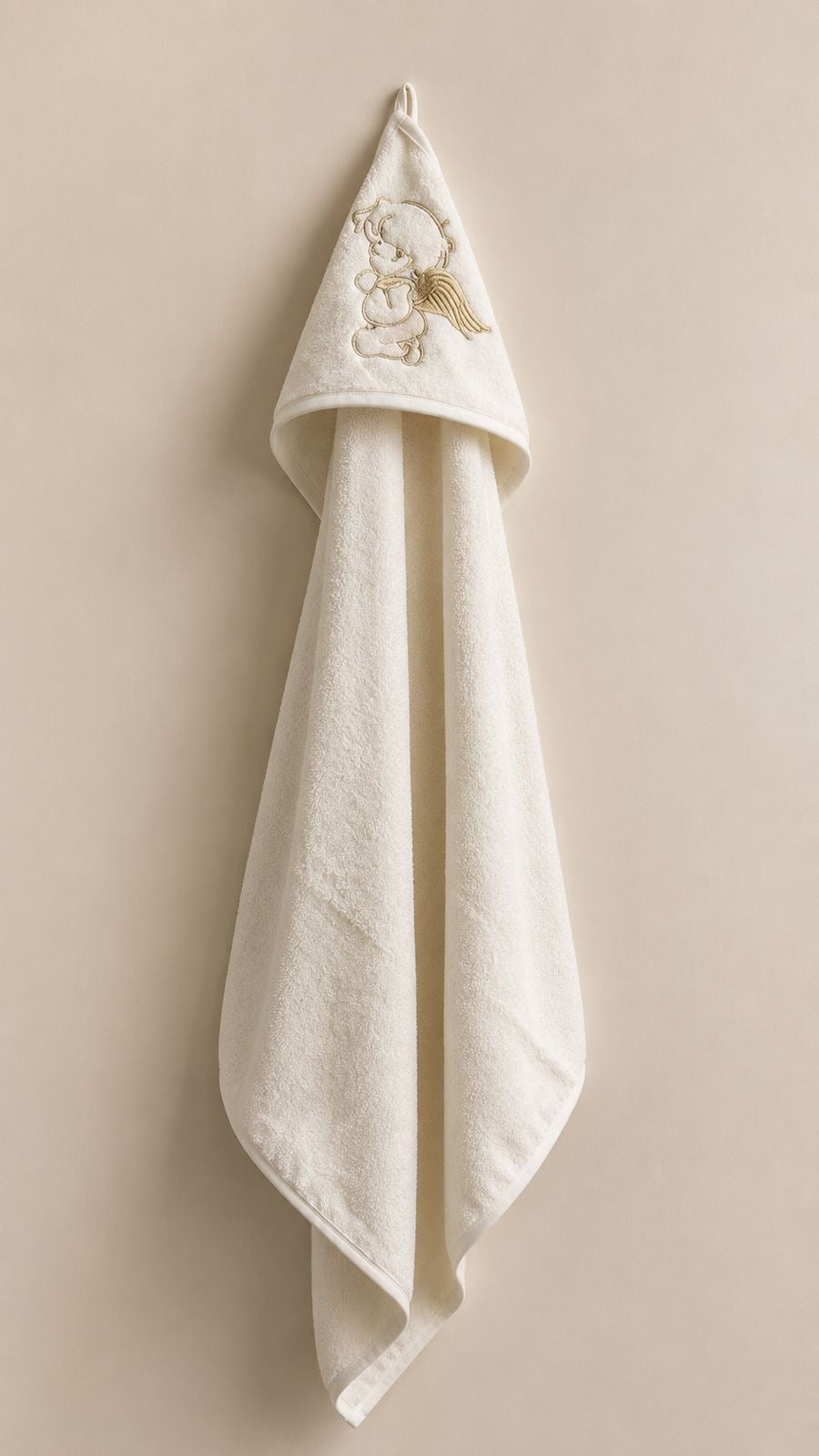 An off-white cotton baby hooded towel with a gold embroidered angel design on the hood, hanging against a solid beige background.