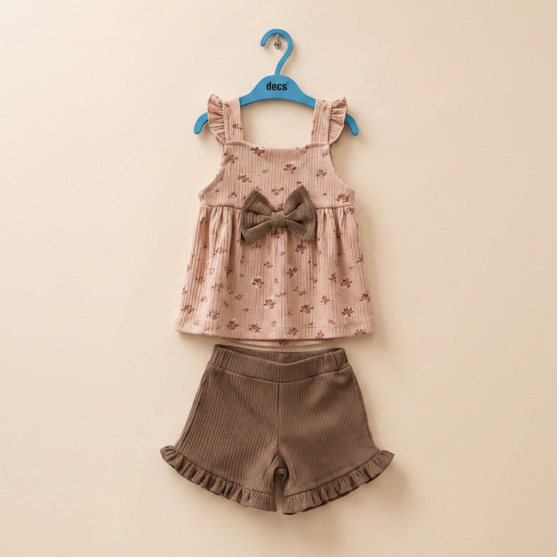 A two-piece toddler girl's outfit set hanging on a blue hanger against a beige wall. The set includes a light pink ribbed sleeveless top with a small floral print, a brown bow on the chest, and matching brown corduroy shorts with a ruffled hem.