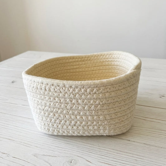 A small woven rope basket in a warm cream color, placed on a light-colored wooden tabletop against a neutral off-white background.