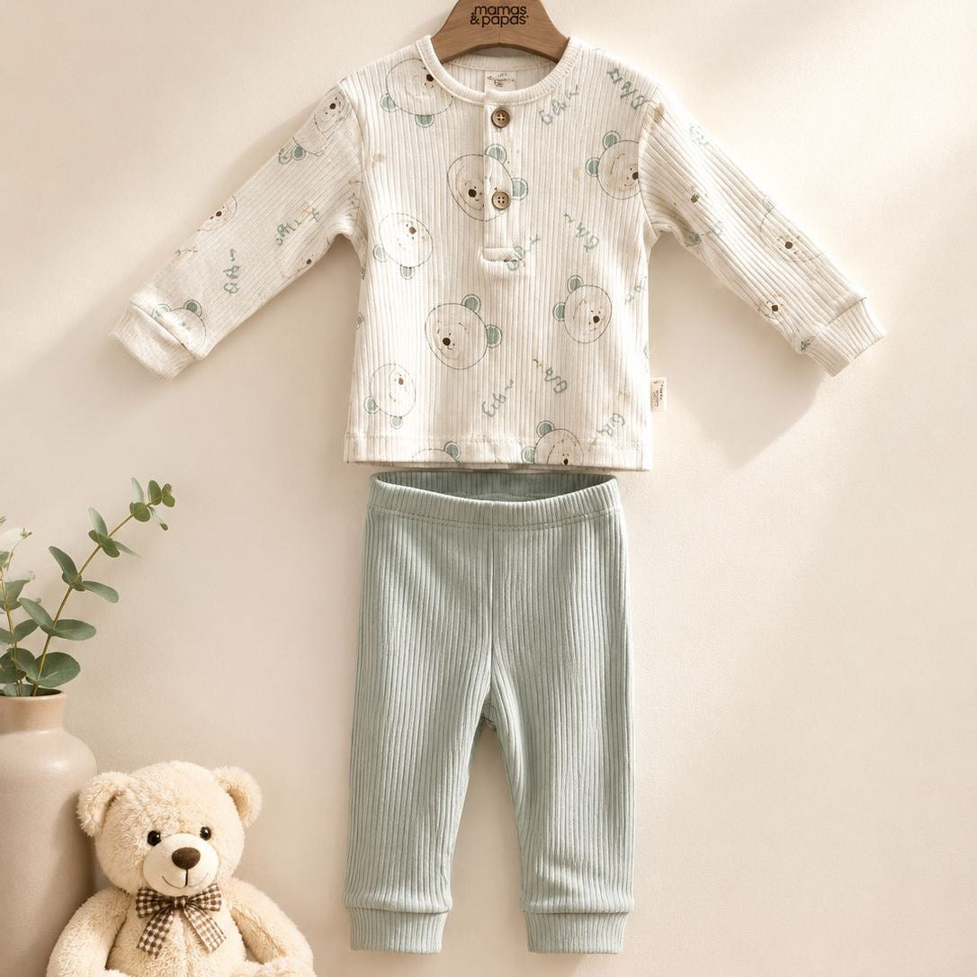A baby two-piece outfit set hanging on a wooden hanger against a warm beige wall. The set includes a long-sleeve button-front top in cream-colored textured fabric with a light blue-gray bear print, and matching light blue-gray ribbed pants.