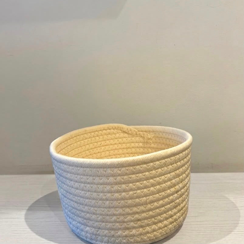 A small round woven rope basket in a warm cream or off-white color, sitting on a light-colored surface against a neutral wall.