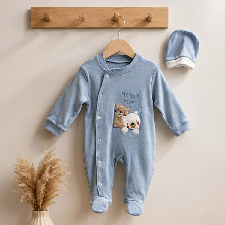 A baby long-sleeve footed romper in light blue hangs on a wooden hanger against a beige wall, with a matching blue bonnet to the right. The romper has a teddy bear and white bear graphic on the chest with the text 'MY FIRST BFF'.