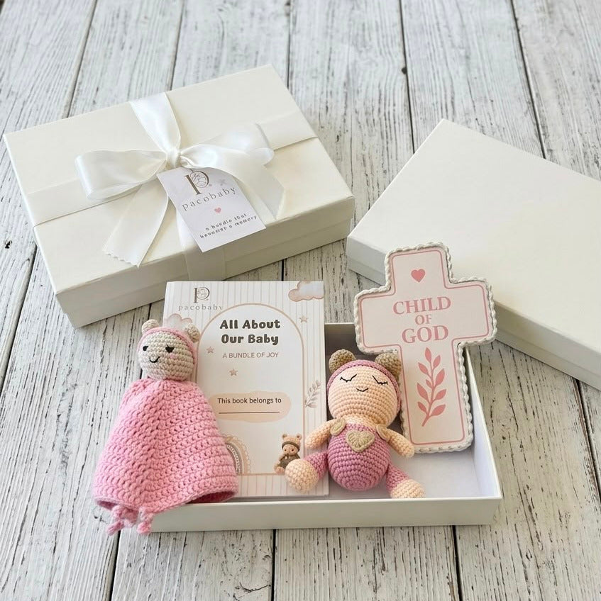 A baby gift set displayed in an open cream-colored box on a whitewashed wooden surface, alongside a closed matching gift box tied with a white ribbon. The set includes a pink crocheted security blanket, a small plush toy in pink and beige, a book titled 'All About Our Baby', and a wooden cross plaque that reads 'CHILD OF GOD'.
