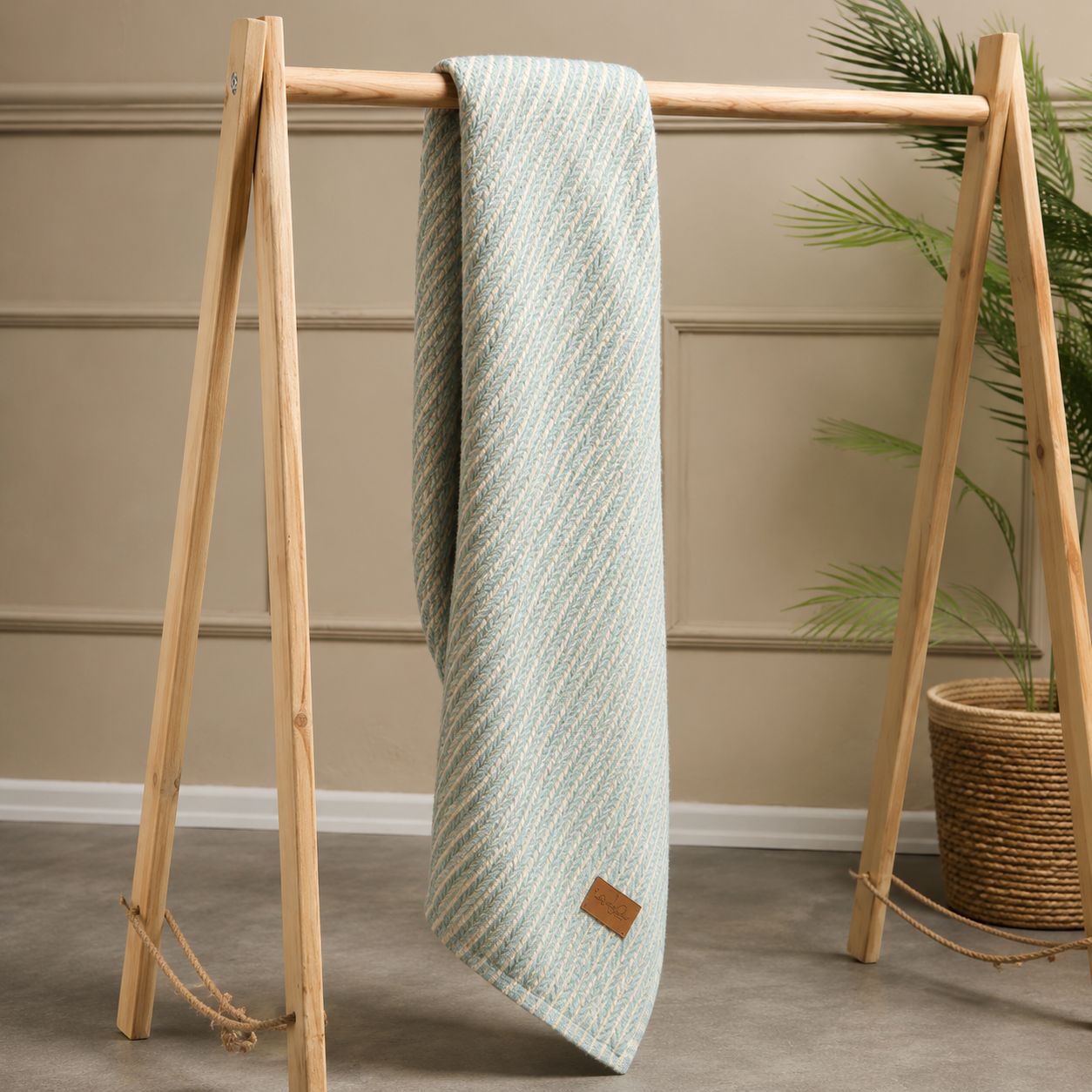 A light sage green textured throw blanket with a diagonal woven pattern, draped over a wooden A-frame stand in a styled room setting with a potted plant.