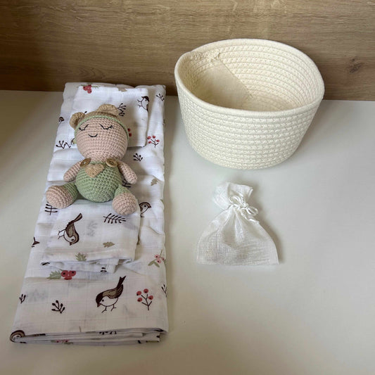 A baby gift set featuring a folded white muslin blanket with a woodland bird pattern and a small crocheted teddy bear, next to a cream woven basket and a small white drawstring pouch on a white surface.