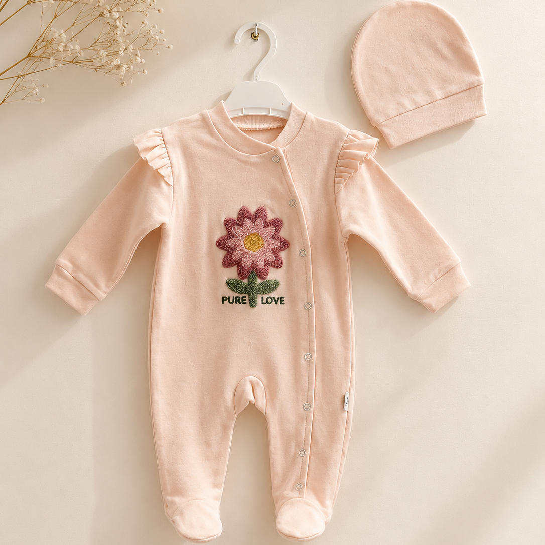 A baby long-sleeve footed romper in pale blush pink, hanging on a white hanger next to a matching knit bonnet. The romper features a colorful embroidered flower on the chest with the text 'PURE LOVE' underneath, set against a warm cream background with dried floral accents in the top-left corner.
