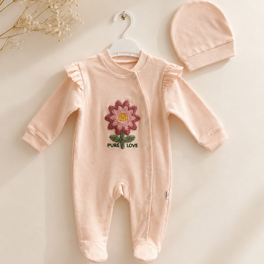 A baby long-sleeve footed romper in pale blush pink, hanging on a white hanger next to a matching knit bonnet. The romper features a colorful embroidered flower on the chest with the text 'PURE LOVE' underneath, set against a warm cream background with dried floral accents in the top-left corner.