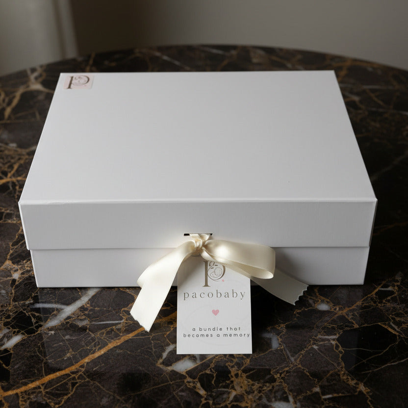 A white rectangular gift box tied with a white satin ribbon and a small hanging tag that reads 'pacobaby', placed on a white surface with a light wood background.