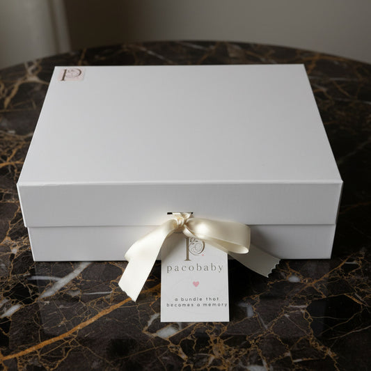 A white rectangular gift box tied with a white satin ribbon and a small hanging tag that reads 'pacobaby', placed on a white surface with a light wood background.