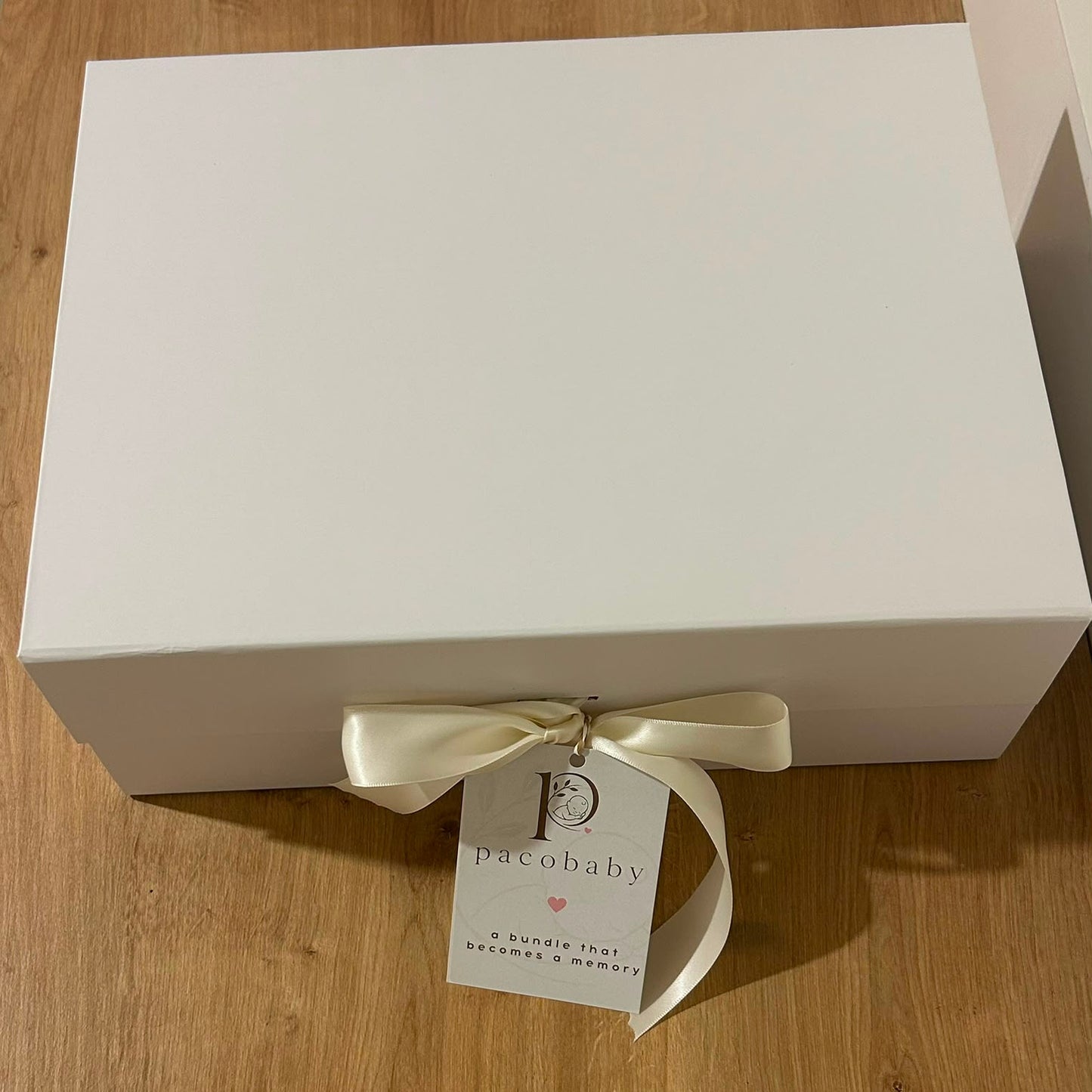 A white rectangular gift box tied with a cream satin ribbon and a hanging tag that reads 'pacobaby', placed on a light wood floor.