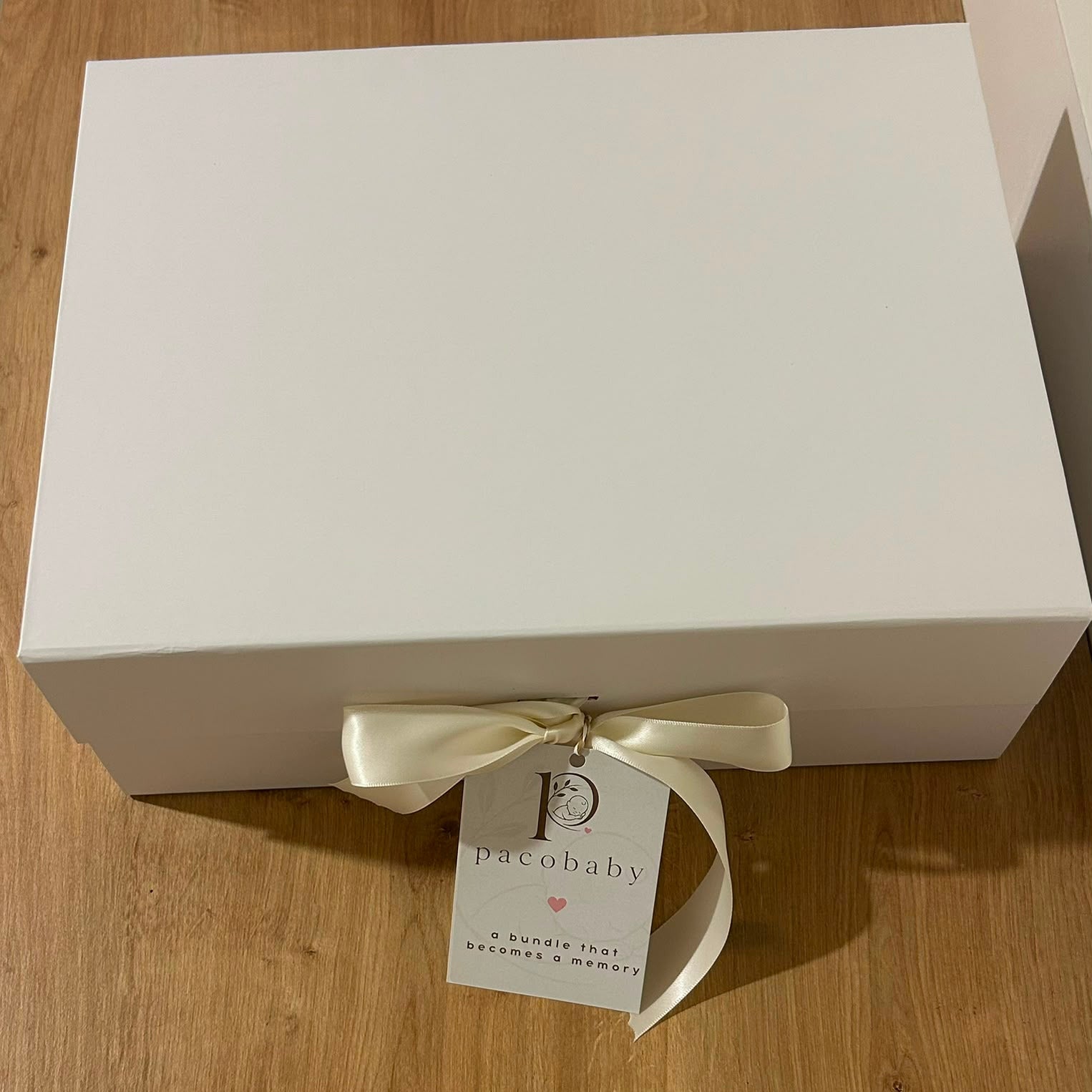 A white rectangular gift box tied with a cream satin ribbon and a hanging tag that reads 'pacobaby', placed on a light wood floor.