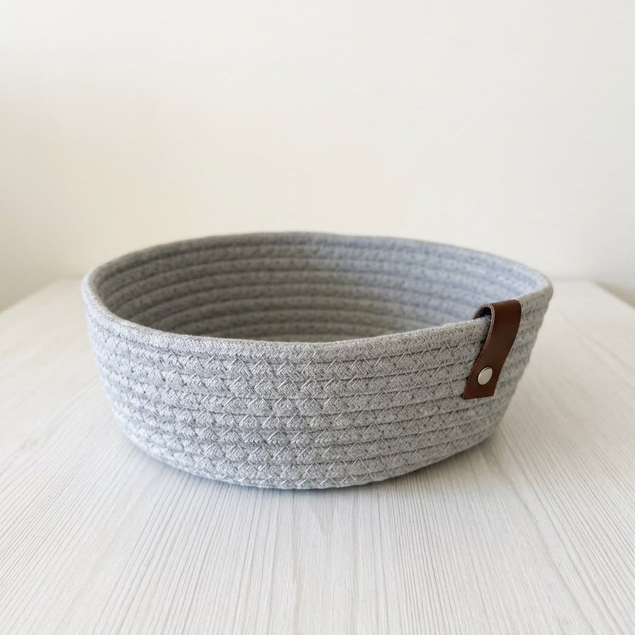 A light gray woven rope storage basket with a brown leather accent tab on the side, resting on a white wooden surface against a plain off-white background.
