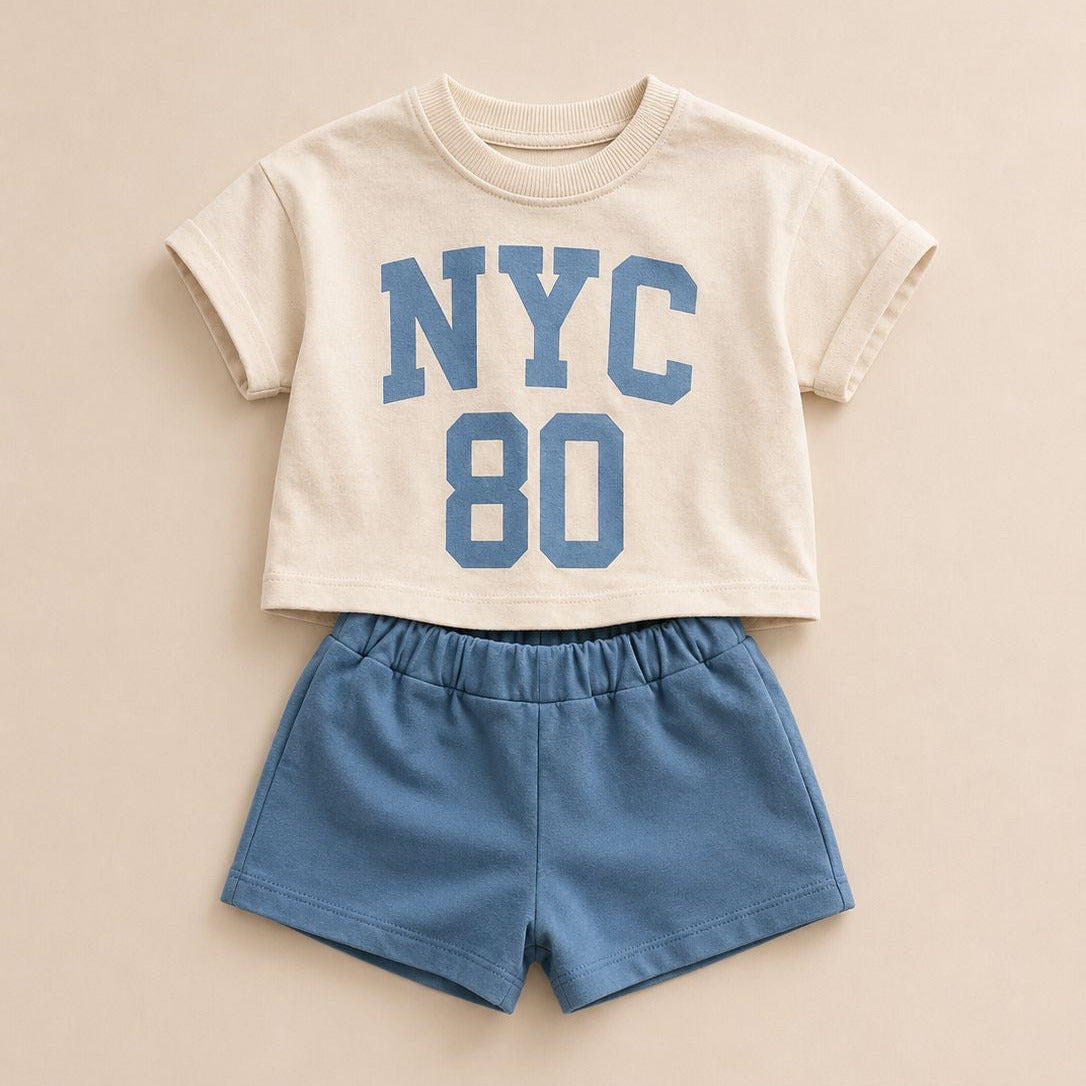A toddler boy's two-piece outfit featuring a cream short-sleeve crop top with blue 'NYC 80' lettering, paired with matching blue shorts, laid flat on a beige background.