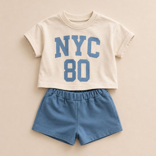 A toddler boy's two-piece outfit featuring a cream short-sleeve crop top with blue 'NYC 80' lettering, paired with matching blue shorts, laid flat on a beige background.