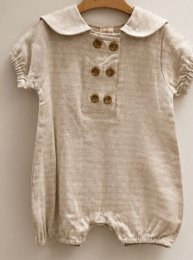 A baby romper in a light beige linen-like fabric, featuring a Peter Pan collar, short sleeves, and a front button closure with brown buttons, hanging on a wooden hanger against a neutral background.