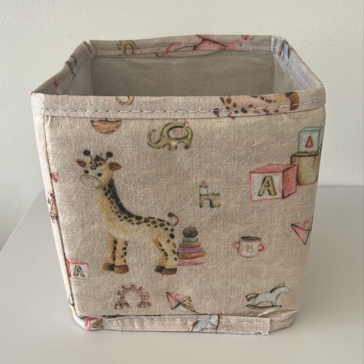 A square fabric storage basket with a safari and children's toy print on a light beige linen-like fabric, set against a plain off-white background.