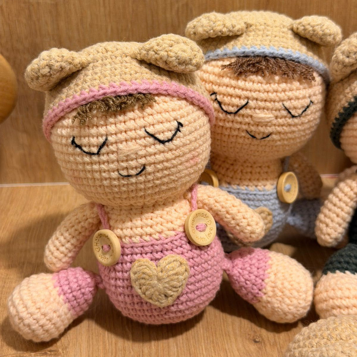 A close-up of handmade crocheted doll toys sitting on a wooden shelf. The main doll in the foreground is beige with pink details, including a pink headband and pink pants with a beige heart motif.
