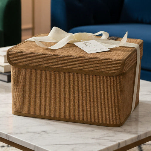 A rectangular woven storage box with a hinged lid, made from natural tan rattan or wicker, tied with a cream-colored ribbon and a paper tag that reads 'pacoubaby'.