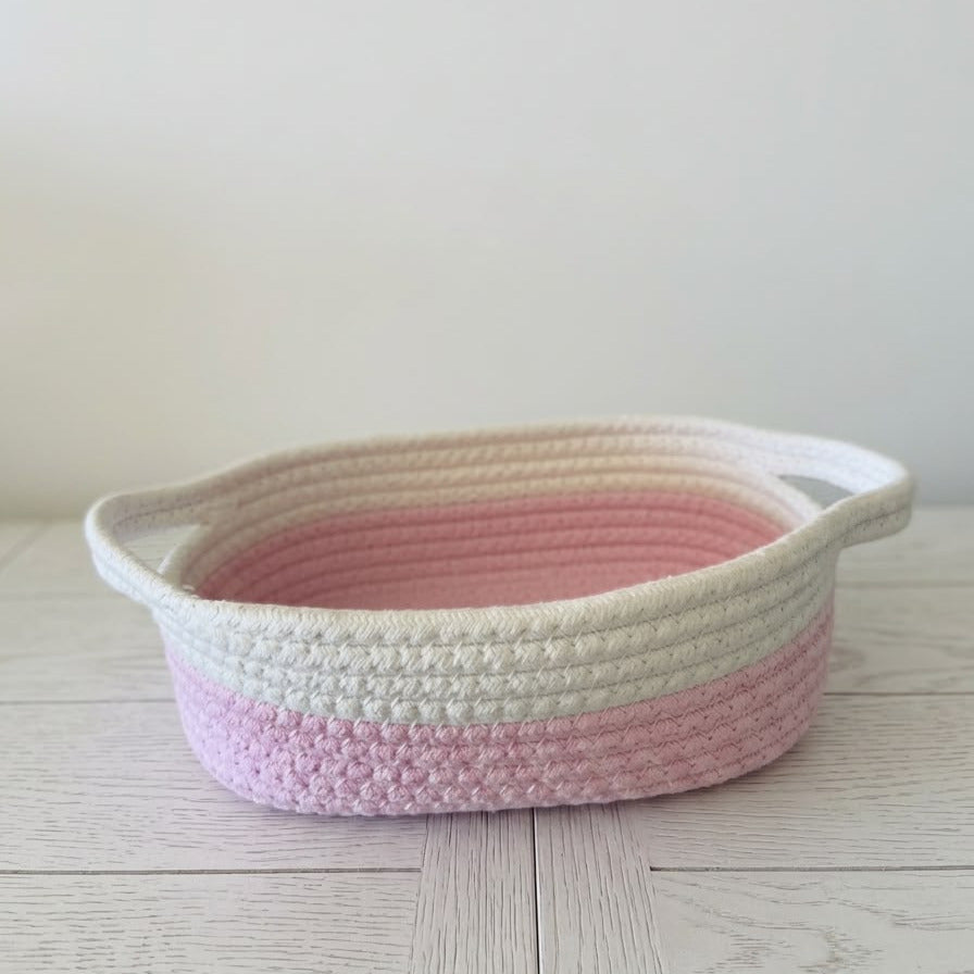 A small crocheted storage basket with two side handles, featuring horizontal stripes of cream, light pink, and a darker pink, placed on a light wood surface against a plain off-white wall.