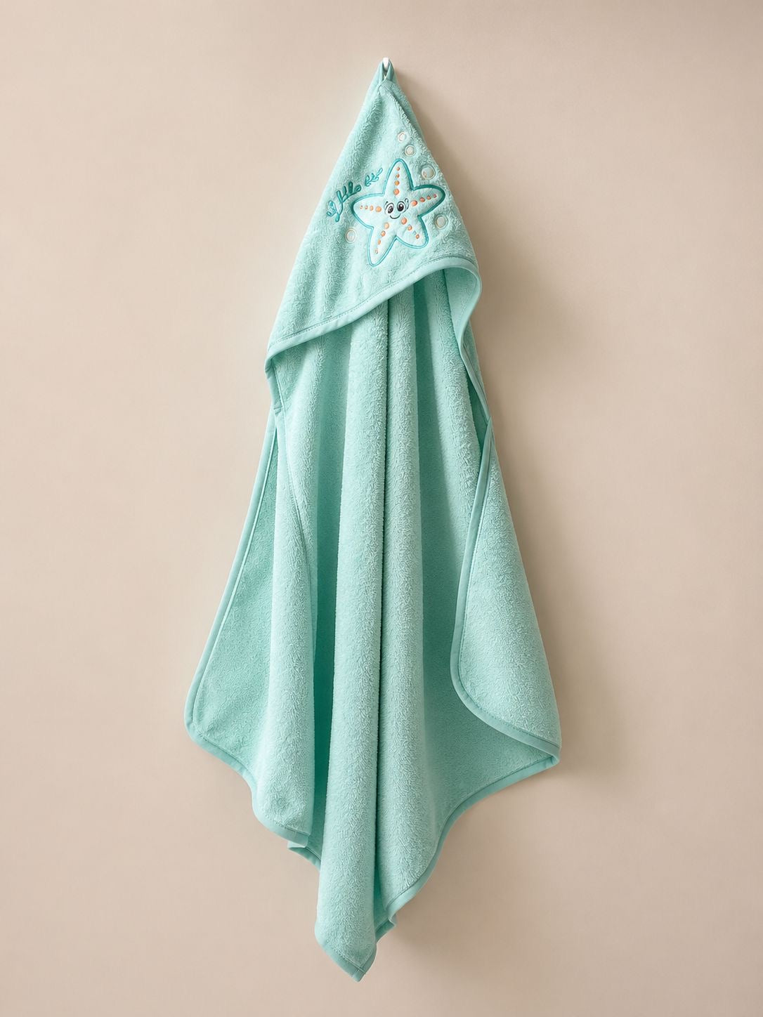 A mint green hooded bath towel with a starfish embroidery on the hood, hanging against a solid beige background.