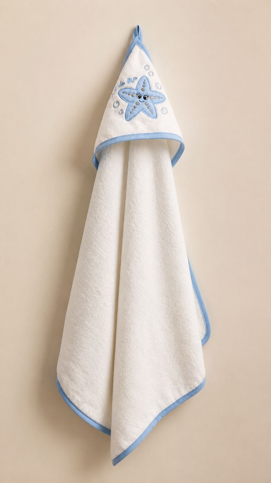 A white hooded baby towel with light blue trim and a blue starfish design on the hood, hanging against a solid beige background.