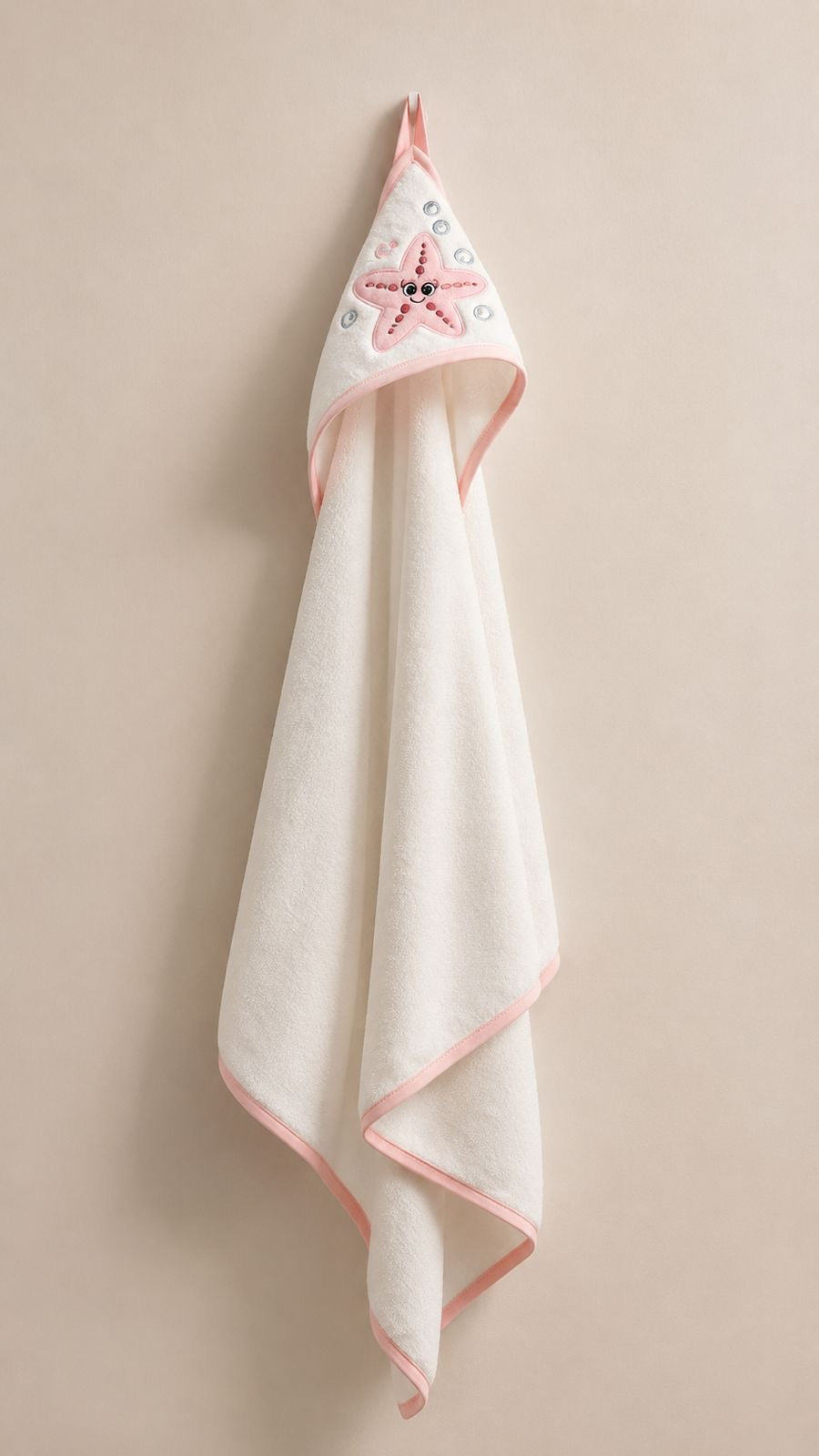 A white hooded baby towel with pink trim and a pink starfish design on the hood, hanging against a solid beige background.