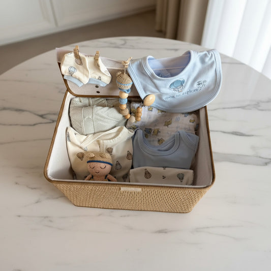 A baby gift set arranged inside an open woven suitcase on a soft cream fabric surface. The set includes folded baby clothes in light blue and cream with small printed motifs, a pale blue bib with embroidered text, a cream knit hat, a wooden bead rattle, and a small plush doll.