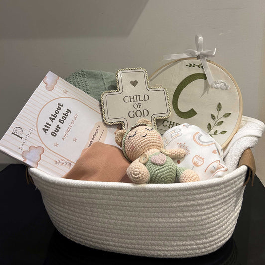 A baby gift basket set in a white woven basket, containing a crocheted cross sign that reads 'CHILD OF GOD', a small plush bear, a swaddle blanket, a folded pink cloth, and a printed card.