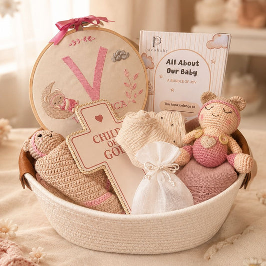A baby shower gift basket filled with soft pink and cream baby-themed items, including a crocheted doll, a plush toy, a book titled 'All About Our Baby,' a wooden cross sign, and a round embroidered piece with a pink letter V.