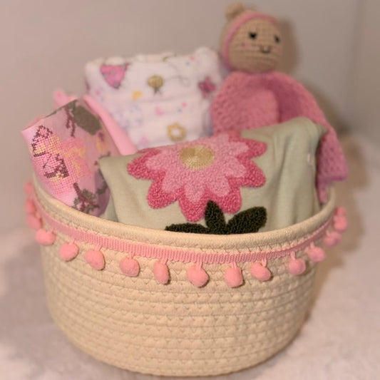 A cream woven basket with pink pom-pom trim holding baby items, including a pink flower applique cloth, a pink patterned fabric piece, a white patterned cloth, and a pink knitted bear toy, against a soft neutral background.