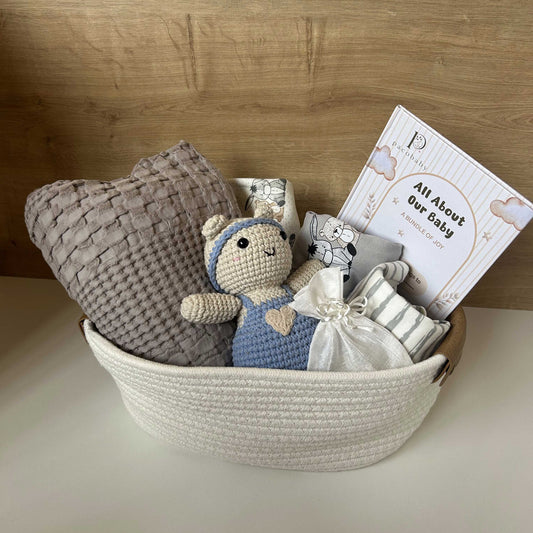 A baby gift basket set in a white woven basket, featuring a gray textured blanket, a crocheted teddy bear in blue and cream, a small white drawstring bag, and a printed card that reads 'All About Our Baby'.