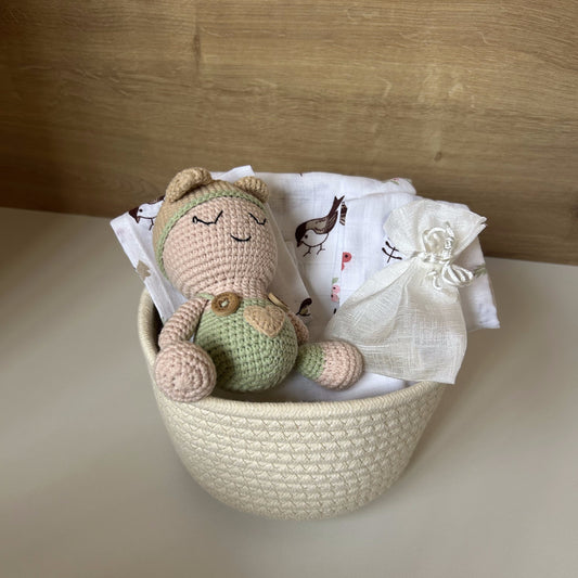 A crocheted baby bear toy in beige and light green sits in a cream woven basket lined with white muslin baby cloths, set against a light wood background.